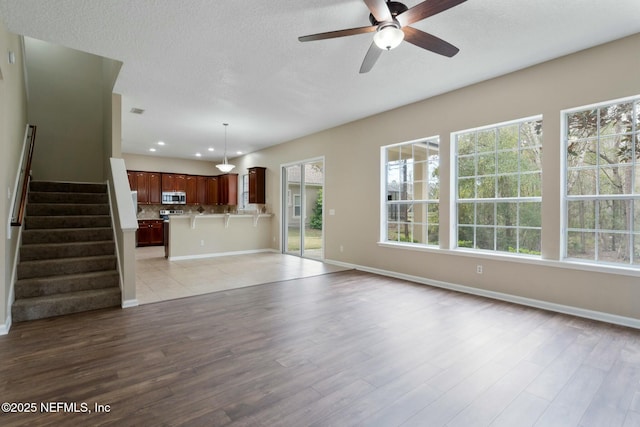 unfurnished living room with a ceiling fan, a textured ceiling, light wood-type flooring, baseboards, and stairs