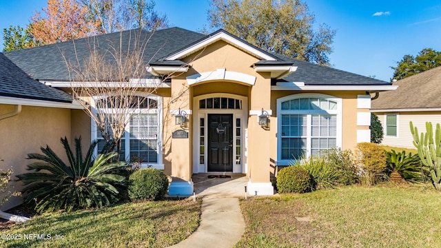 view of exterior entry with roof with shingles, a yard, and stucco siding