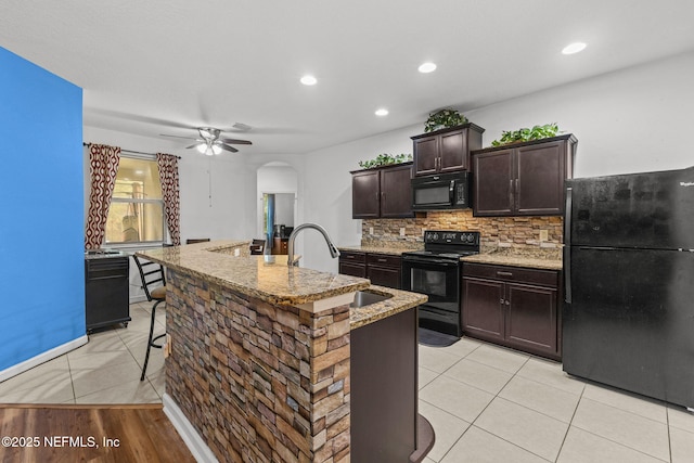 kitchen with arched walkways, light stone counters, a kitchen breakfast bar, black appliances, and backsplash