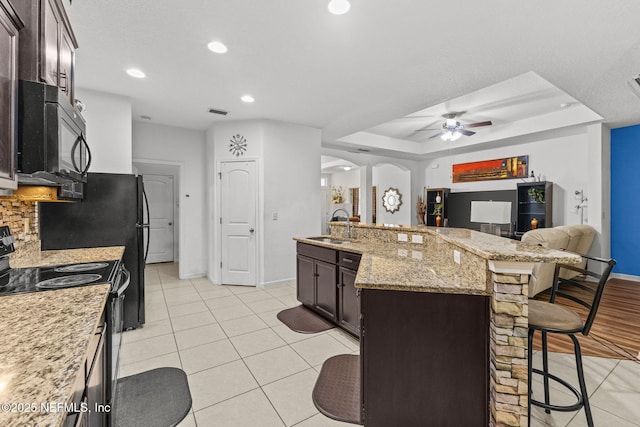 kitchen featuring arched walkways, a breakfast bar area, dark brown cabinets, black appliances, and a sink