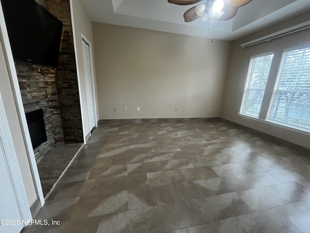 spare room with a ceiling fan, a raised ceiling, a stone fireplace, and baseboards