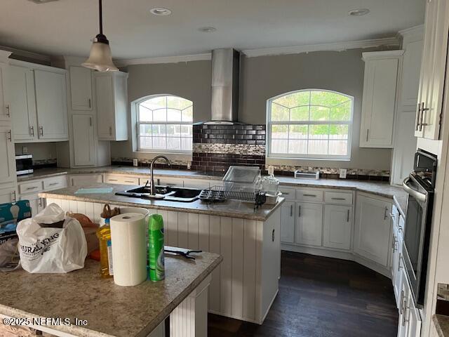 kitchen with wall chimney range hood, a healthy amount of sunlight, a center island with sink, white cabinetry, and black oven