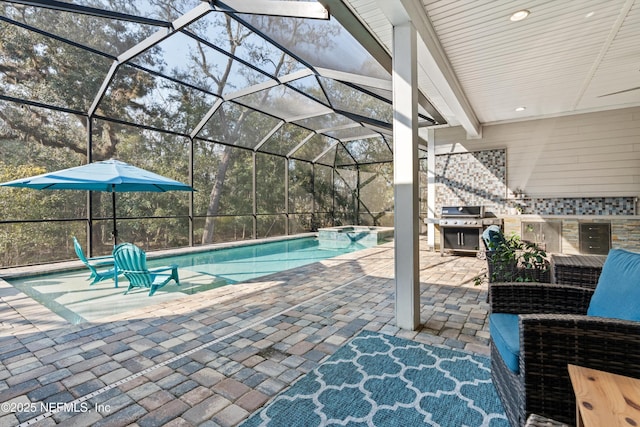 view of pool featuring glass enclosure, a pool with connected hot tub, an outdoor kitchen, and a patio