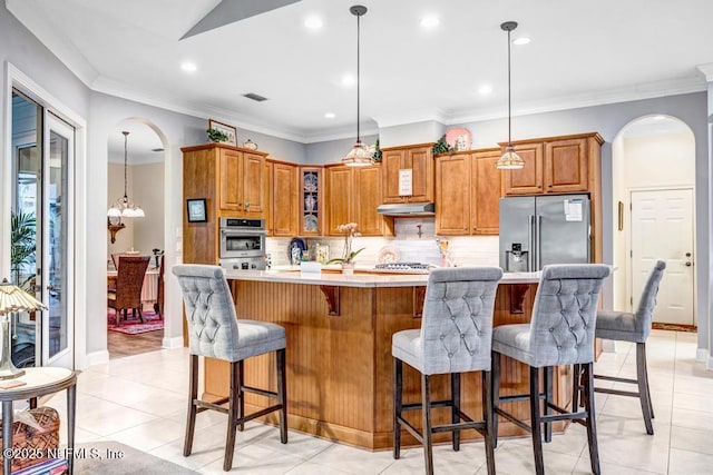 kitchen with arched walkways, stainless steel appliances, brown cabinetry, and under cabinet range hood