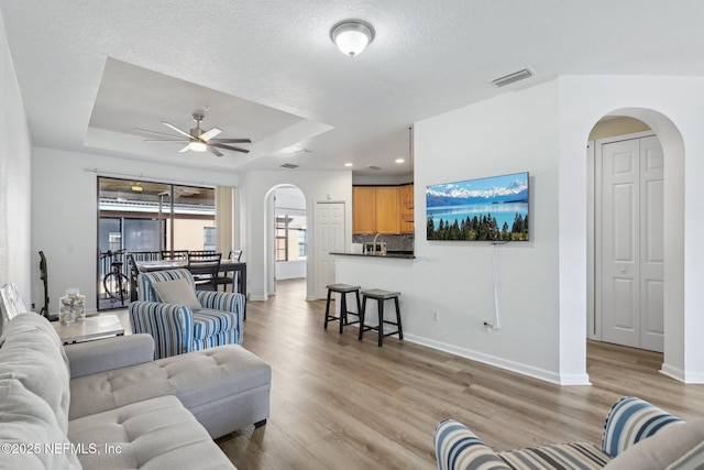 living room with arched walkways, a tray ceiling, light wood-style flooring, and visible vents