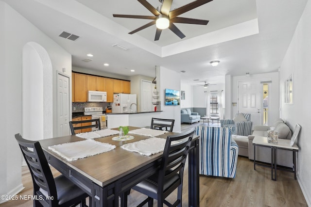 dining area with arched walkways, light wood finished floors, a raised ceiling, visible vents, and a ceiling fan