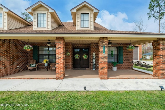 view of exterior entry featuring a ceiling fan, covered porch, brick siding, and roof with shingles