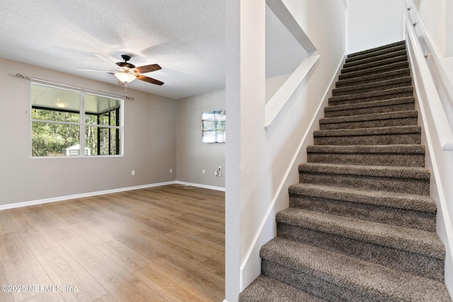 stairway with a ceiling fan, a textured ceiling, baseboards, and wood finished floors