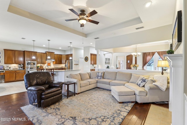 living room featuring a tray ceiling, light wood-style flooring, ceiling fan with notable chandelier, and visible vents