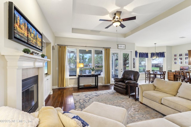 living area featuring a tiled fireplace, a tray ceiling, baseboards, ceiling fan, and dark wood-style flooring