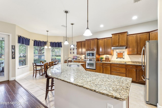 kitchen featuring a kitchen bar, brown cabinets, a sink, under cabinet range hood, and appliances with stainless steel finishes