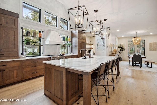 kitchen with a notable chandelier, light wood finished floors, custom exhaust hood, hanging light fixtures, and a large island with sink