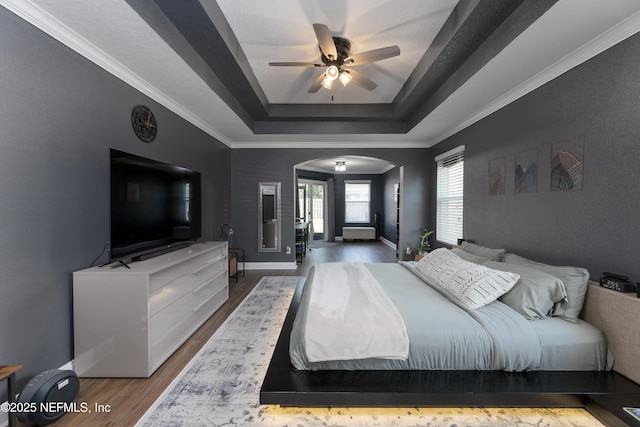 bedroom featuring arched walkways, a tray ceiling, crown molding, wood finished floors, and baseboards