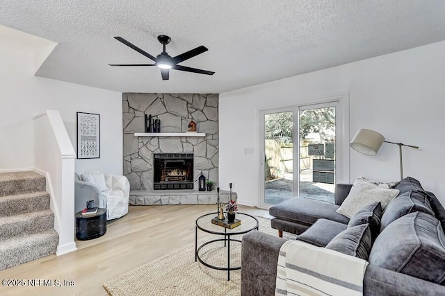 living room featuring ceiling fan, a stone fireplace, a textured ceiling, wood finished floors, and stairs