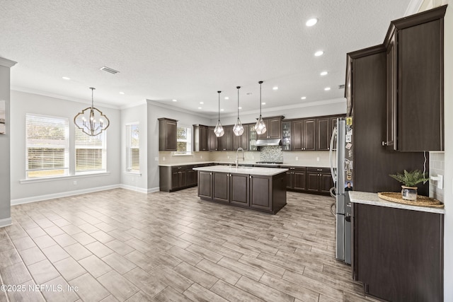 kitchen featuring tasteful backsplash, dark brown cabinets, under cabinet range hood, a chandelier, and a center island with sink