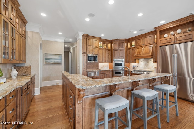 kitchen featuring brown cabinetry, ornamental molding, light stone countertops, stainless steel appliances, and light wood-type flooring