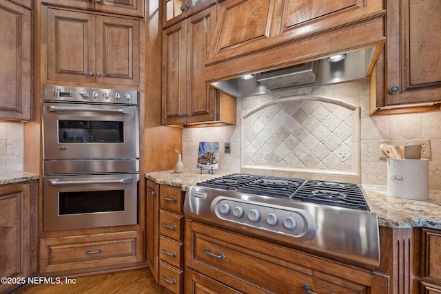 kitchen with stainless steel appliances, premium range hood, brown cabinetry, and backsplash