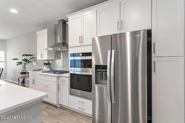 kitchen with wall chimney exhaust hood, appliances with stainless steel finishes, light countertops, white cabinetry, and backsplash