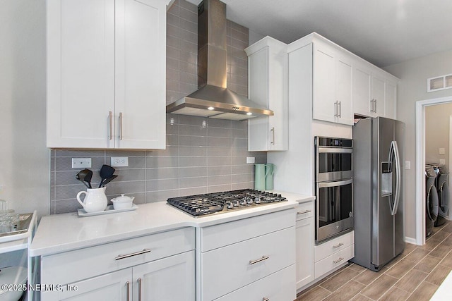 kitchen featuring stainless steel appliances, decorative backsplash, washing machine and dryer, white cabinetry, and wall chimney range hood