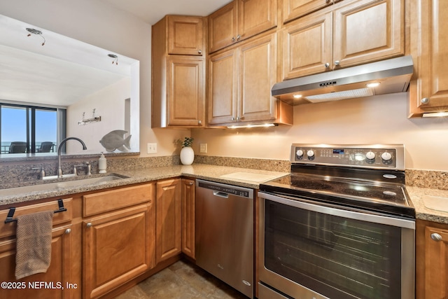 kitchen with under cabinet range hood, light stone counters, appliances with stainless steel finishes, brown cabinetry, and a sink