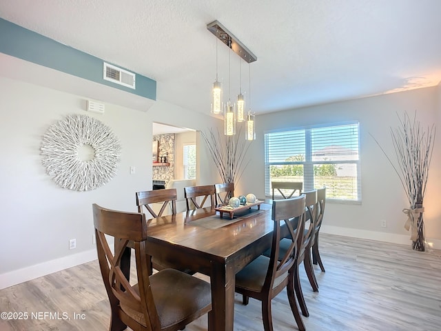 dining room with a textured ceiling, a stone fireplace, visible vents, baseboards, and light wood finished floors