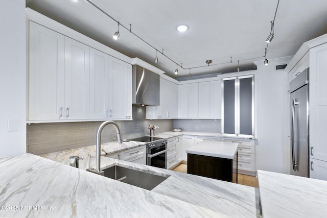 kitchen with a sink, wall chimney range hood, appliances with stainless steel finishes, backsplash, and light stone countertops