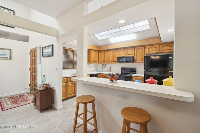 kitchen featuring a skylight, light countertops, brown cabinets, black appliances, and a kitchen bar