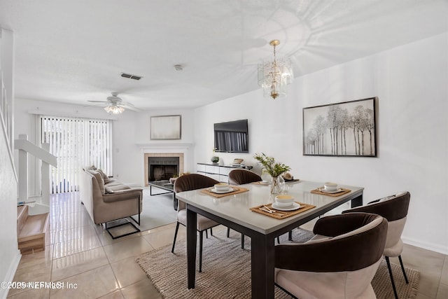 dining space featuring visible vents, a tiled fireplace, ceiling fan with notable chandelier, light tile patterned flooring, and baseboards