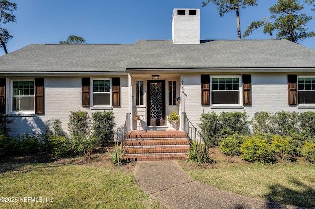 view of front facade featuring a front yard, brick siding, a chimney, and roof with shingles