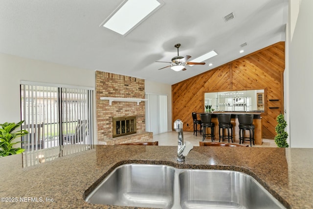 kitchen with lofted ceiling, wooden walls, a sink, a brick fireplace, and dark stone countertops