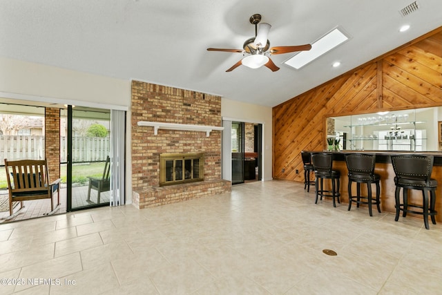 living room featuring a fireplace, visible vents, a ceiling fan, wood walls, and vaulted ceiling with skylight