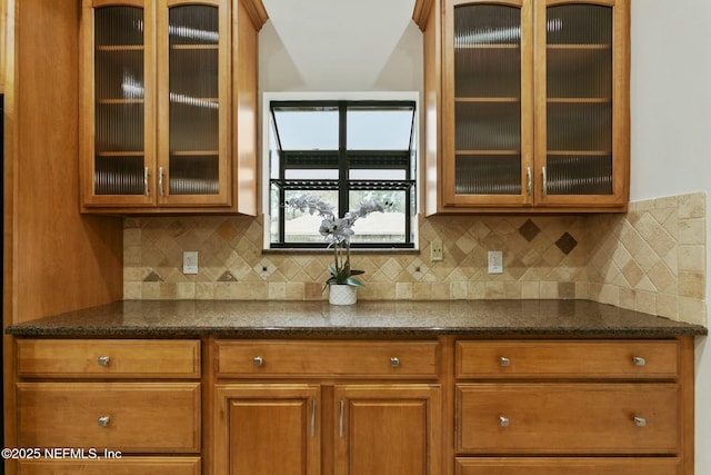 kitchen featuring brown cabinetry, dark stone countertops, backsplash, and glass insert cabinets