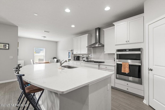 kitchen with a center island with sink, wall chimney exhaust hood, double oven, gas cooktop, and a sink