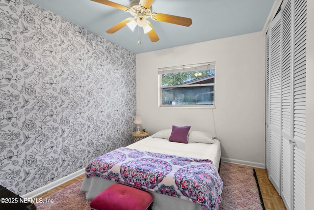 bedroom with a closet, an accent wall, a ceiling fan, baseboards, and wallpapered walls