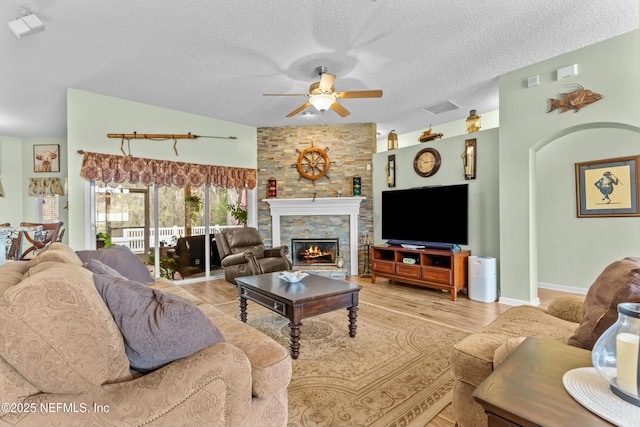 living room featuring a textured ceiling, a stone fireplace, visible vents, a ceiling fan, and light wood-type flooring