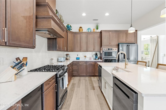 kitchen with light stone counters, stainless steel appliances, backsplash, a kitchen island with sink, and premium range hood