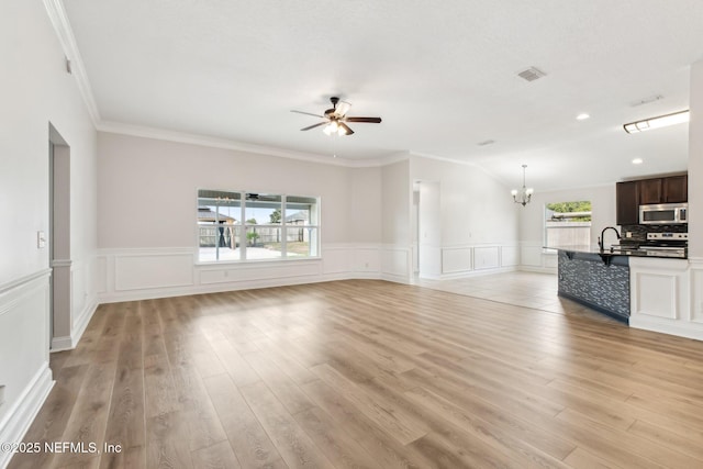 unfurnished living room featuring a wainscoted wall, light wood-style floors, ornamental molding, a sink, and ceiling fan with notable chandelier