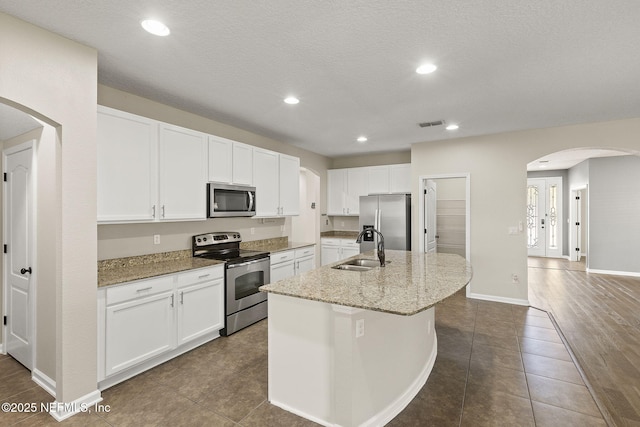 kitchen with a center island with sink, stainless steel appliances, arched walkways, white cabinetry, and a sink