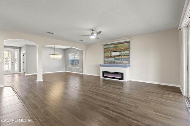 unfurnished living room featuring visible vents, a textured ceiling, a glass covered fireplace, dark wood finished floors, and arched walkways