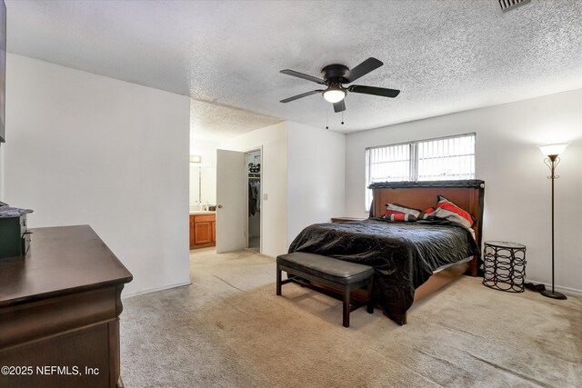 bedroom featuring a walk in closet, light carpet, ensuite bathroom, a ceiling fan, and a textured ceiling