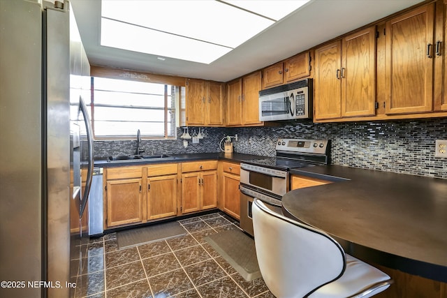 kitchen featuring brown cabinets, a sink, backsplash, dark countertops, and appliances with stainless steel finishes