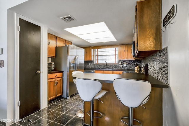 kitchen with visible vents, a sink, dark countertops, stainless steel fridge, and brown cabinetry