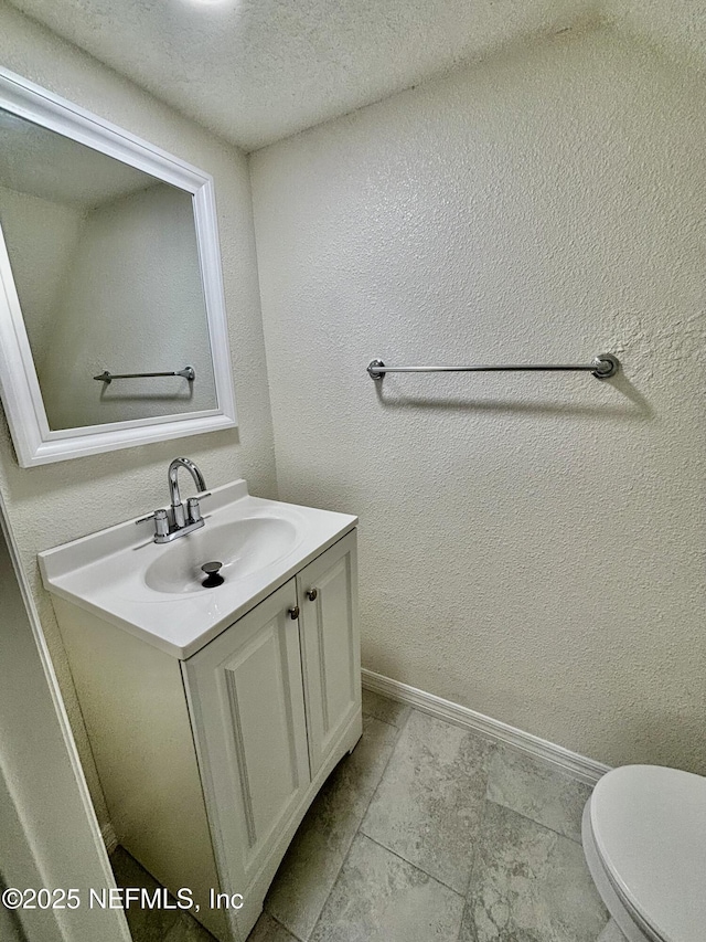 half bathroom featuring toilet, a textured wall, vanity, and baseboards