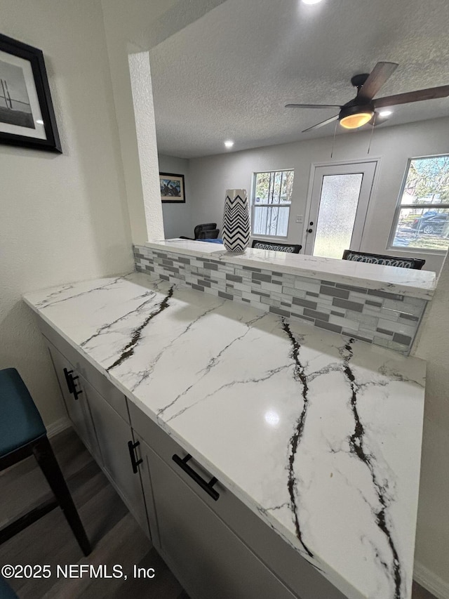 kitchen featuring a ceiling fan, a peninsula, light stone countertops, a textured ceiling, and backsplash