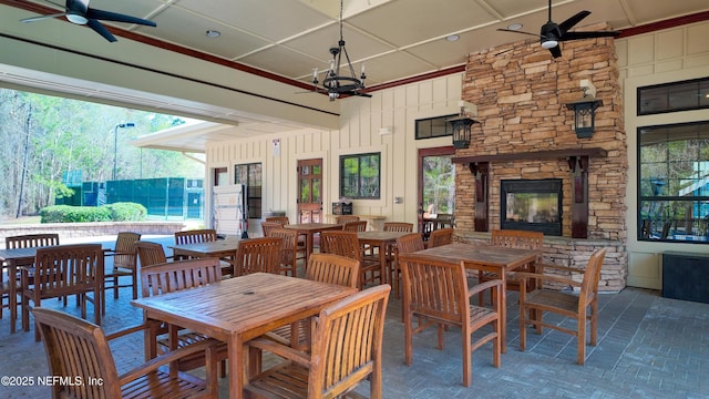 dining space with brick floor, an outdoor stone fireplace, and ceiling fan with notable chandelier