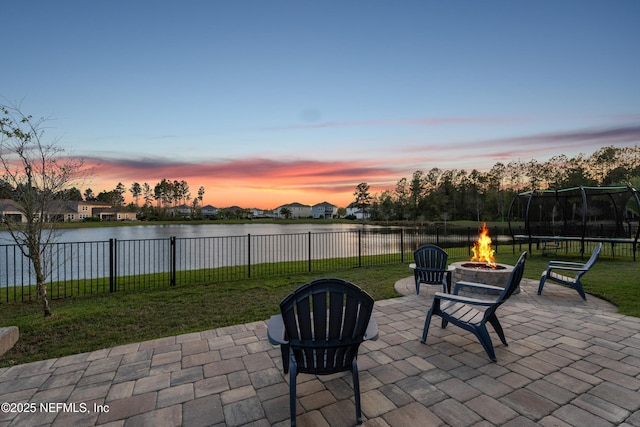 patio terrace at dusk featuring an outdoor fire pit, a fenced backyard, a water view, a trampoline, and a lawn