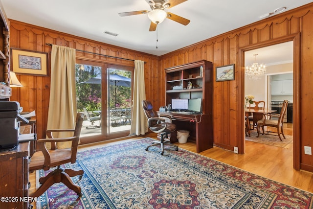 home office with light wood finished floors, visible vents, wood walls, ornamental molding, and ceiling fan with notable chandelier