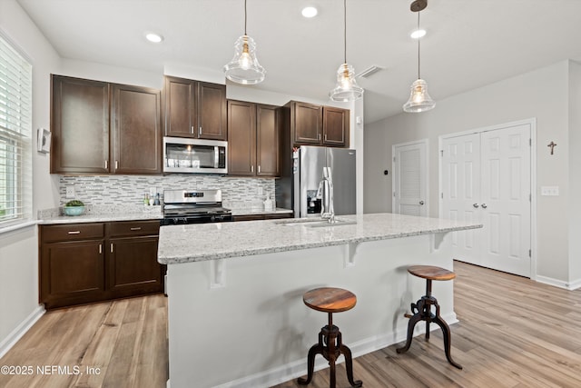 kitchen featuring light wood finished floors, decorative backsplash, appliances with stainless steel finishes, a breakfast bar area, and dark brown cabinets