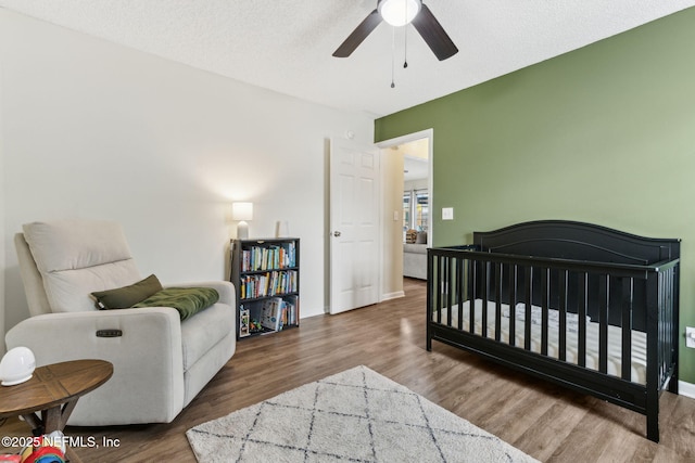 bedroom featuring a textured ceiling, wood finished floors, a nursery area, baseboards, and ceiling fan