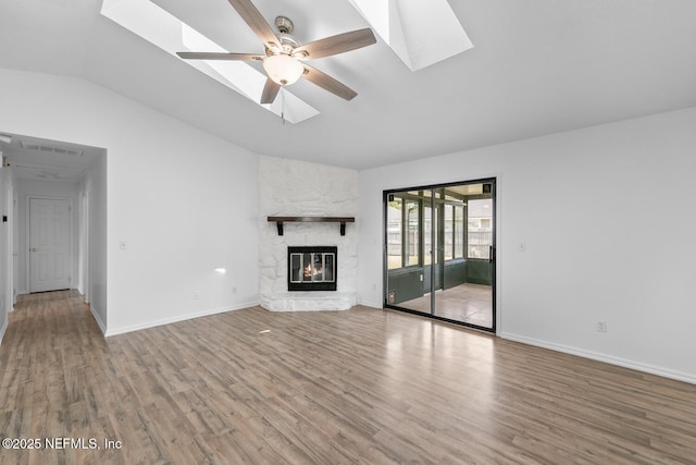 unfurnished living room with a ceiling fan, a stone fireplace, wood finished floors, vaulted ceiling with skylight, and baseboards